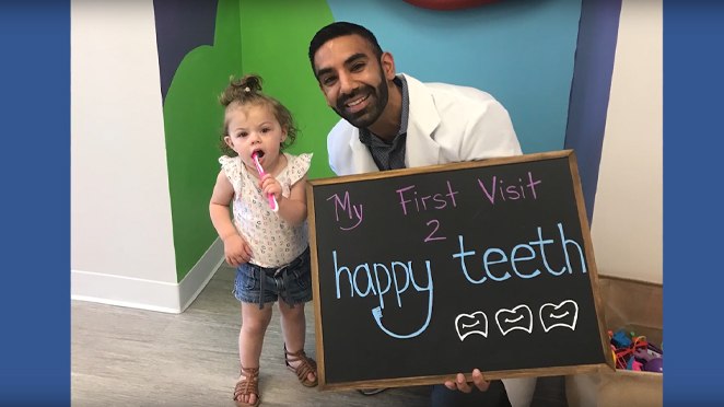 Doctor Singh smiling with toddler patient holding chalk sign reading my first visit 2 Happy Teeth